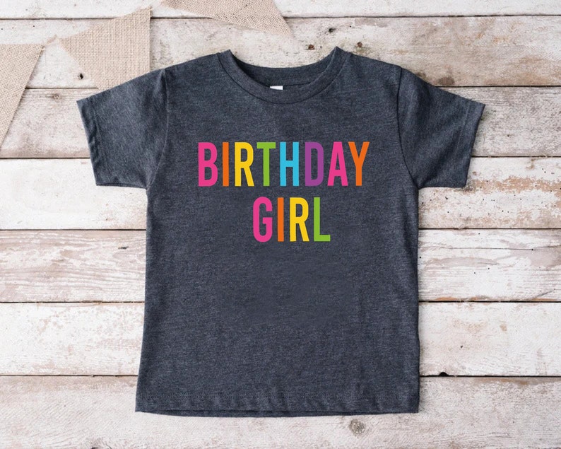 May include: A dark grey t-shirt with the words "BIRTHDAY GIRL" printed in rainbow colors.