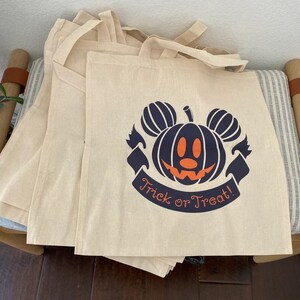 May include: A set of five beige canvas tote bags with black handles. The front of each bag features a black and orange pumpkin with Mickey Mouse ears and the text "Trick or Treat!"
