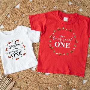 May include: Two white short-sleeved t-shirts with red strawberry designs. The larger shirt says "the berry sweet one" and the smaller shirt says "sister of the berry sweet one".