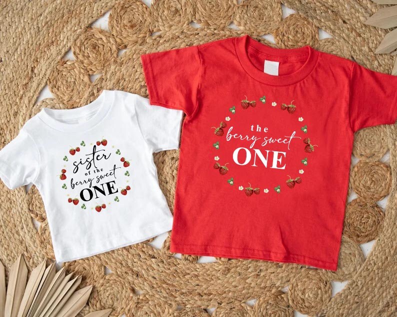 May include: Two white short-sleeved t-shirts with red strawberry designs. The larger shirt says "the berry sweet one" and the smaller shirt says "sister of the berry sweet one".