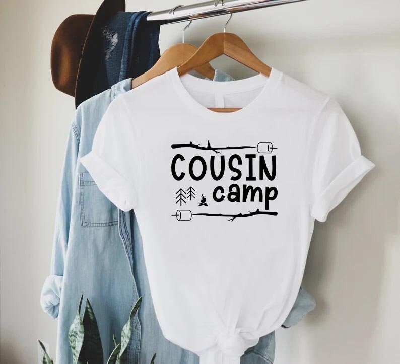May include: White t-shirt with black text that reads "COUSIN camp" with a campfire and pine trees graphic.