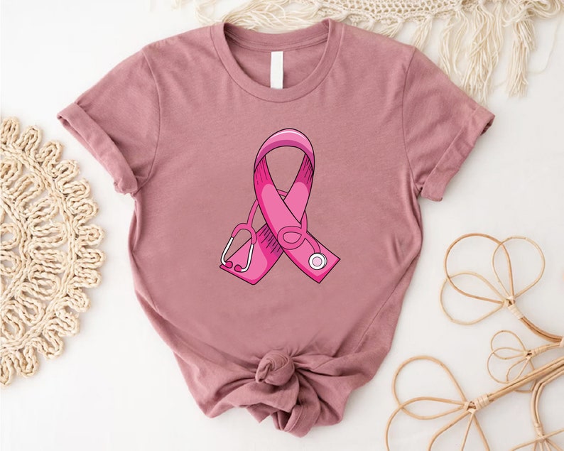 May include: A pink t-shirt with a pink ribbon and a stethoscope printed on the front. The ribbon is a symbol of breast cancer awareness.