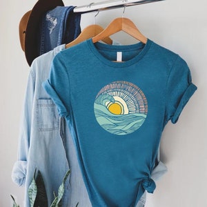 May include: A teal blue t-shirt with a graphic of a yellow sun setting over a blue and green wave. The wave is outlined in black and the sun has a pink and orange glow.
