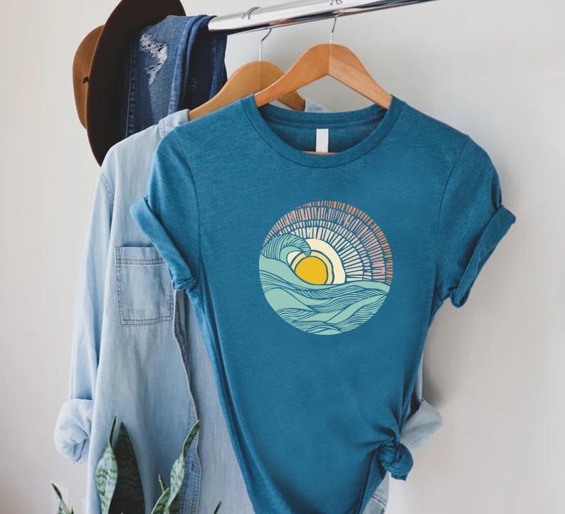 May include: A teal blue t-shirt with a graphic of a yellow sun setting over a blue and green wave. The wave is outlined in black and the sun has a pink and orange glow.