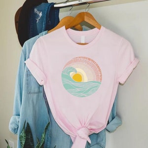 May include: A pink t-shirt with a graphic of a stylized sun setting over a wave. The sun is yellow and the wave is blue and green.