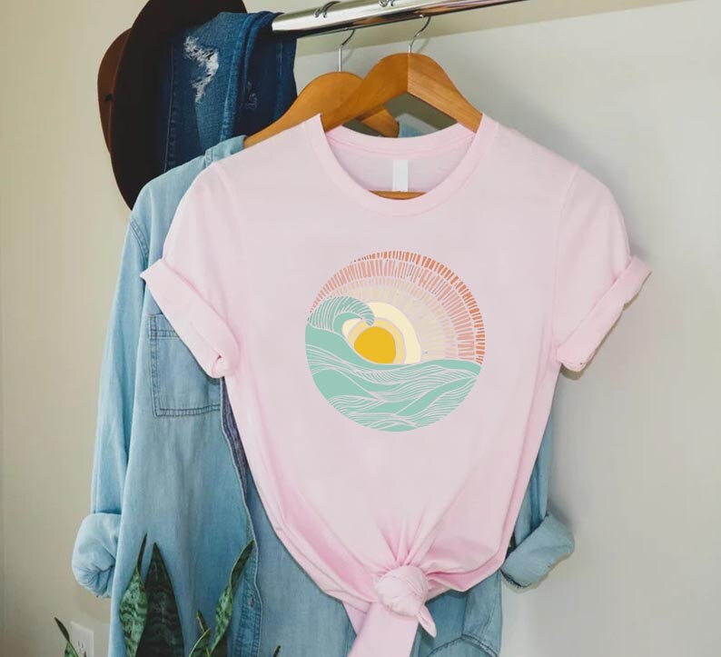May include: A pink t-shirt with a graphic of a stylized sun setting over a wave. The sun is yellow and the wave is blue and green.