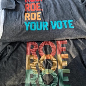 May include: Two gray t-shirts with the text "ROE. ROE. ROE. YOUR VOTE" printed in blue and a rainbow-colored "ROE" printed in red, yellow, and green.