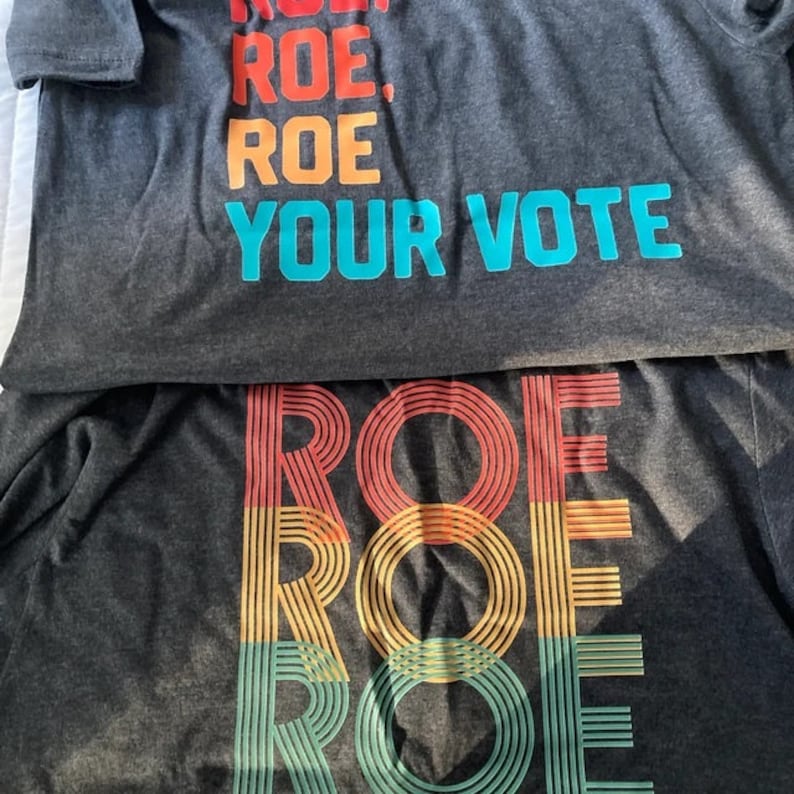 May include: Two gray t-shirts with the text "ROE. ROE. ROE. YOUR VOTE" printed in blue and a rainbow-colored "ROE" printed in red, yellow, and green.
