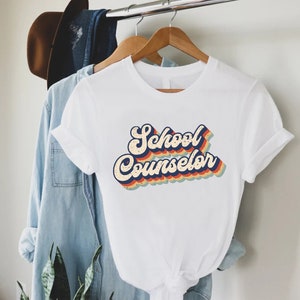 May include: White t-shirt with a retro-style graphic that says "School Counselor" in rainbow colors.
