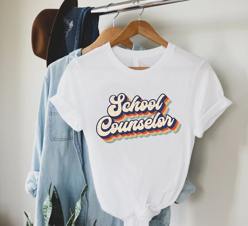 May include: White t-shirt with a retro-style graphic that says "School Counselor" in rainbow colors.