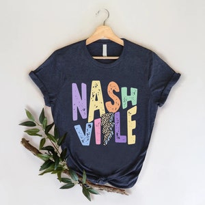 Puede incluir: Una camiseta azul oscuro con un gr&aacute;fico colorido que dice "NASHVILLE" en un estilo retro. Las letras est&aacute;n delineadas en blanco y tienen un aspecto desgastado. El gr&aacute;fico tambi&eacute;n incluye un rayo con un patr&oacute;n de estampado de leopardo.