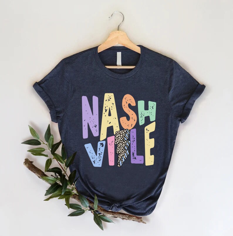 Puede incluir: Una camiseta azul oscuro con un gr&aacute;fico colorido que dice "NASHVILLE" en un estilo retro. Las letras est&aacute;n delineadas en blanco y tienen un aspecto desgastado. El gr&aacute;fico tambi&eacute;n incluye un rayo con un patr&oacute;n de estampado de leopardo.