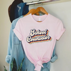 May include: Pink t-shirt with a retro-style graphic that says "School Counselor" in rainbow colors.