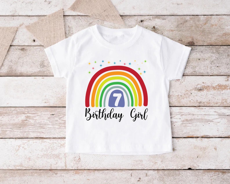 Peut inclure: T-shirt blanc avec un motif arc-en-ciel et le chiffre 7 dans un cercle bleu. Le texte "Birthday Girl" est &eacute;crit sous l'arc-en-ciel.