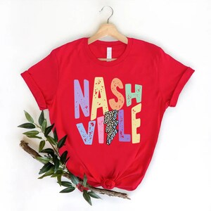 Puede incluir: Camiseta roja con un gr&aacute;fico colorido de "Nashville" que presenta un rayo con un dise&ntilde;o de estampado de leopardo.