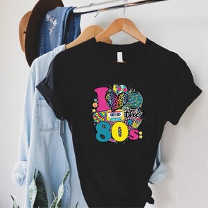 May include: Black t-shirt featuring a graphic print of a heart with a leopard print, a cassette tape, a disco ball, and the text "I love the 80's".