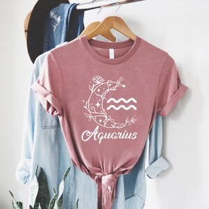 May include: A dusty rose t-shirt with a white graphic of a crescent moon with a floral design and the word "Aquarius" underneath.