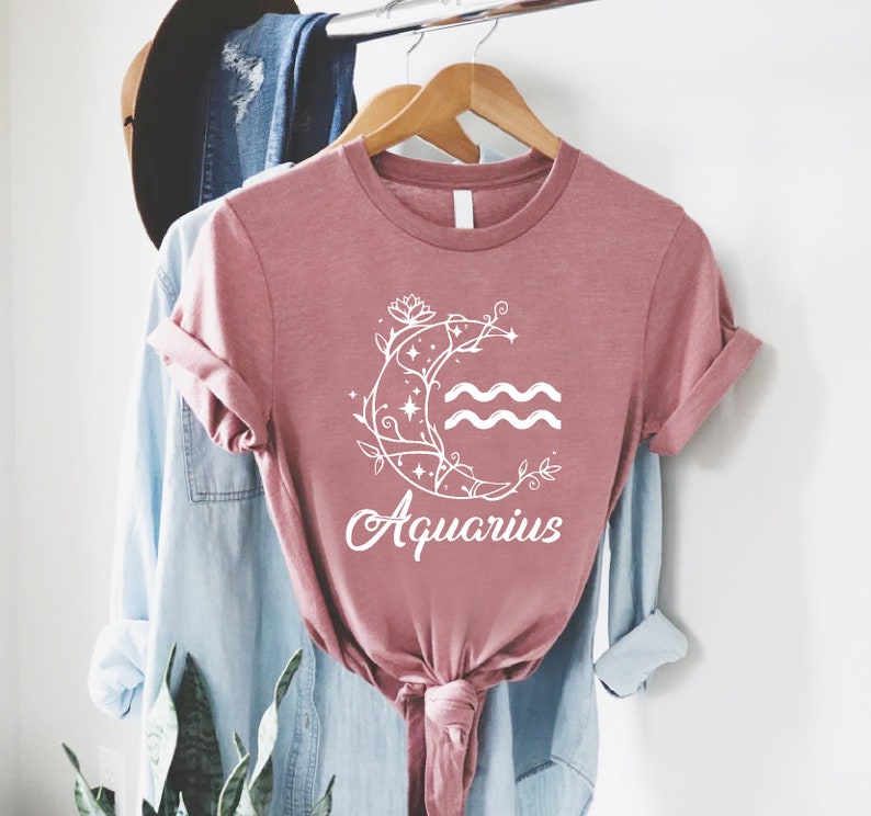 May include: A dusty rose t-shirt with a white graphic of a crescent moon with a floral design and the word "Aquarius" underneath.