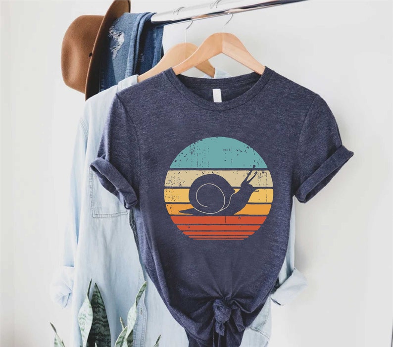 K&ouml;nnte beinhalten: Ein marineblaues T-Shirt mit einer Grafik im Vintage-Stil eines Schnecken in einem farbenfrohen Sonnenuntergang. Die Grafik hat einen abgenutzten Look und verf&uuml;gt &uuml;ber ein Farbschema in T&uuml;rkis, Gelb, Orange und Rot.