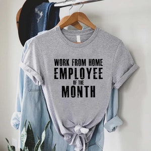 May include: A heather gray t-shirt with the text "WORK FROM HOME EMPLOYEE OF THE MONTH" in black block letters. The shirt is hanging on a wooden hanger, with a denim shirt and a hat in the background.