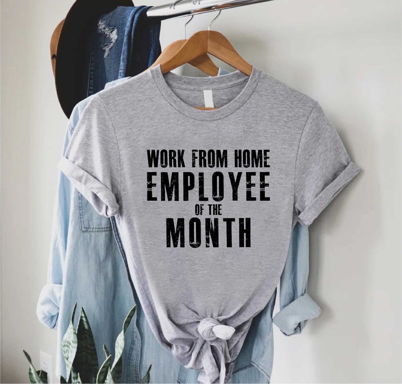 May include: A heather gray t-shirt with the text "WORK FROM HOME EMPLOYEE OF THE MONTH" in black block letters. The shirt is hanging on a wooden hanger, with a denim shirt and a hat in the background.