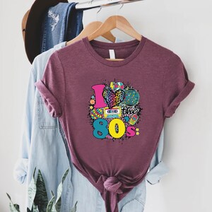 May include: A maroon t-shirt featuring a graphic print of a heart with a leopard print, a disco ball, a cassette tape, a Rubik's cube, and the text "I love the 80s".
