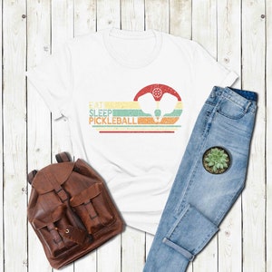 Pu&ograve; includere: Maglietta bianca con una grafica in stile vintage che dice "Eat Sleep Pickleball" con un paio di racchette da pickleball al centro. La grafica &egrave; in tonalit&agrave; di giallo, arancione, verde e blu. La maglietta &egrave; appoggiata su una superficie di legno bianco con uno zaino di pelle marrone e un paio di jeans blu.