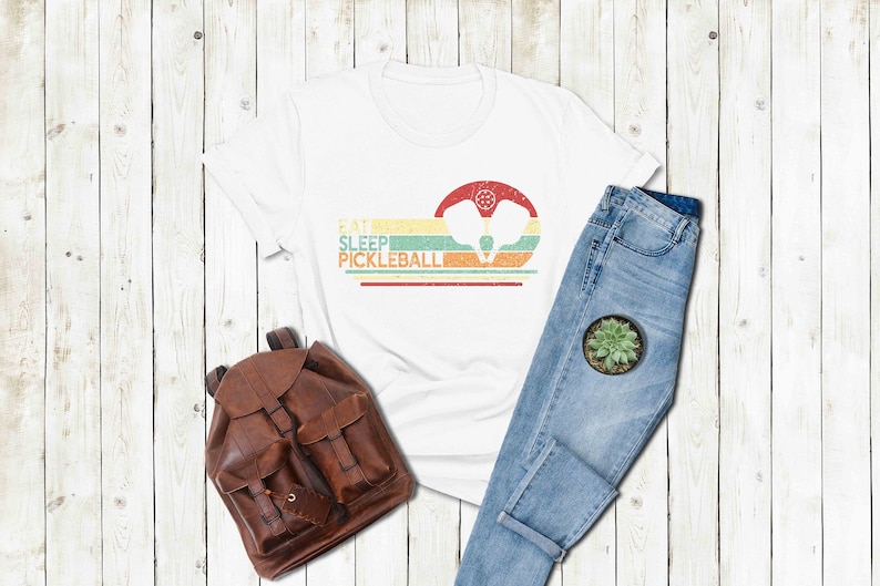 Pu&ograve; includere: Maglietta bianca con una grafica in stile vintage che dice "Eat Sleep Pickleball" con un paio di racchette da pickleball al centro. La grafica &egrave; in tonalit&agrave; di giallo, arancione, verde e blu. La maglietta &egrave; appoggiata su una superficie di legno bianco con uno zaino di pelle marrone e un paio di jeans blu.