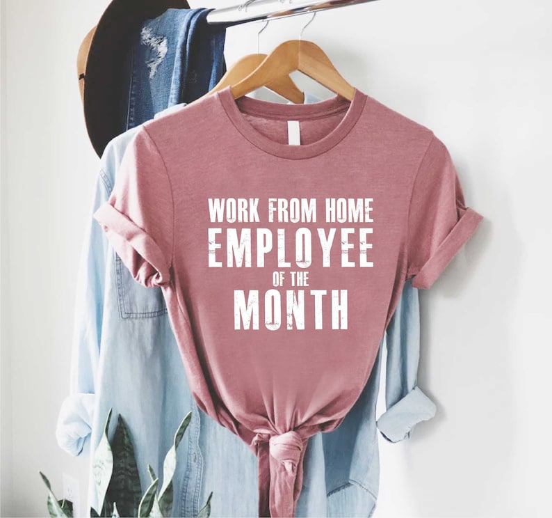 May include: A dusty rose t-shirt with the text "WORK FROM HOME EMPLOYEE OF THE MONTH" in white block letters. The shirt is knotted at the waist and hangs on a wooden hanger with other clothing items in the background.