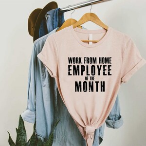 May include: A light pink t-shirt with the text "WORK FROM HOME EMPLOYEE OF THE MONTH" in black. The shirt is hanging on a wooden hanger with a denim shirt and a brown hat in the background.