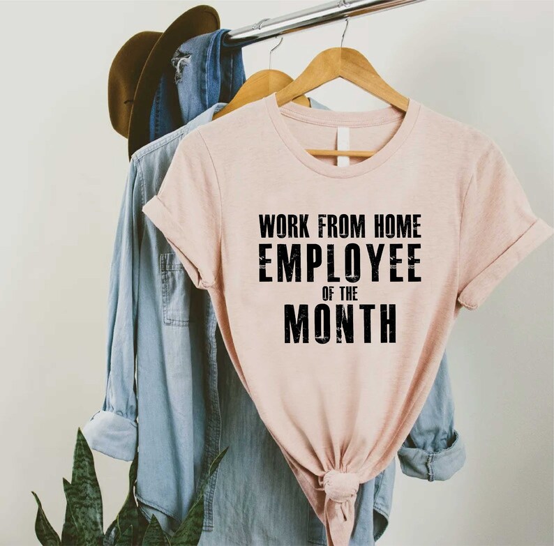 May include: A light pink t-shirt with the text "WORK FROM HOME EMPLOYEE OF THE MONTH" in black. The shirt is hanging on a wooden hanger with a denim shirt and a brown hat in the background.