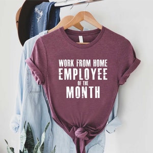 May include: A maroon t-shirt with rolled-up sleeves and the text "WORK FROM HOME EMPLOYEE OF THE MONTH" in white block letters. The shirt is hanging on a wooden hanger with other clothing items in the background.