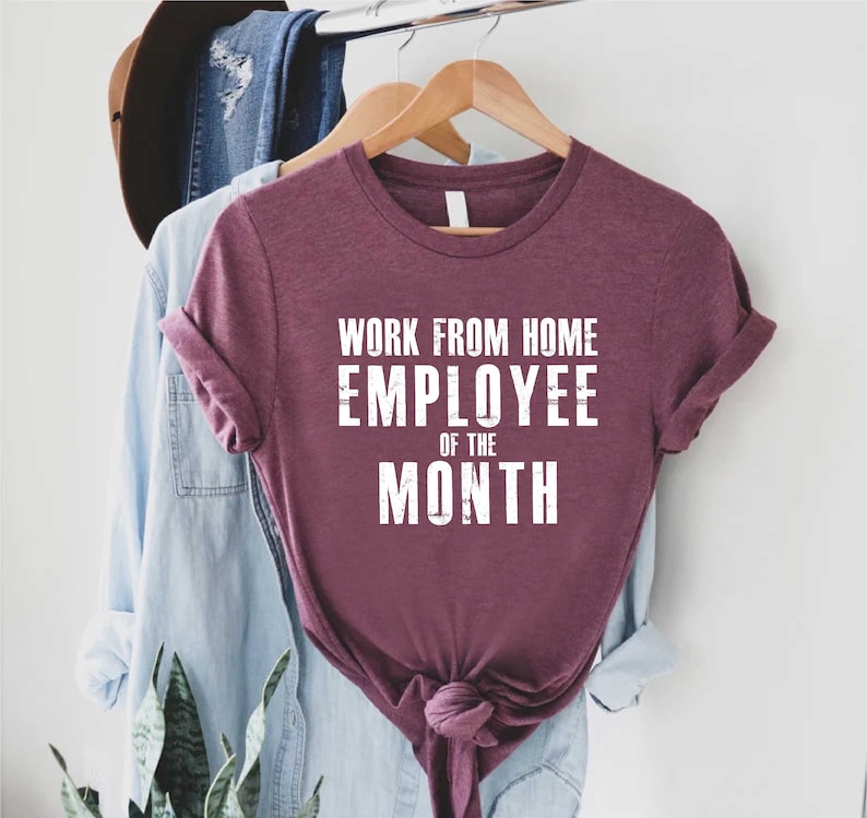 May include: A maroon t-shirt with rolled-up sleeves and the text "WORK FROM HOME EMPLOYEE OF THE MONTH" in white block letters. The shirt is hanging on a wooden hanger with other clothing items in the background.