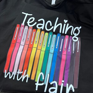 May include: Black t-shirt with the text "Teaching with flair" in white. A row of colorful markers are displayed below the text.