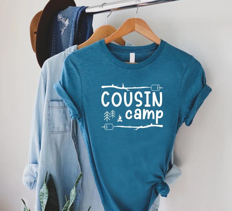 May include: A teal blue t-shirt with white text that reads "COUSIN camp" with a campfire, trees, and marshmallows graphic.