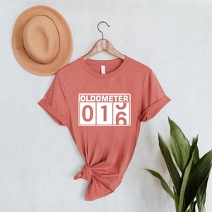 May include: A coral coloured t-shirt with a white graphic that says "Oldometer 016".