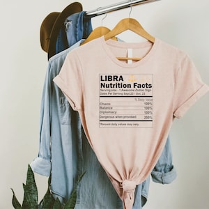 May include: A pink t-shirt with a graphic of a nutrition facts label for Libra, a zodiac sign. The label lists the following traits: Charm, Balance, Diplomacy, and Dangerous when provoked. The text "Libra Nutrition Facts" is at the top of the label, and the text "Serving size - 1 Awesome Zodiac Sign" is below it.
