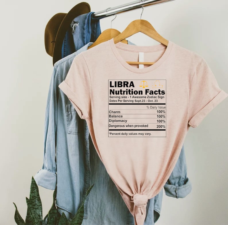 May include: A pink t-shirt with a graphic of a nutrition facts label for Libra, a zodiac sign. The label lists the following traits: Charm, Balance, Diplomacy, and Dangerous when provoked. The text "Libra Nutrition Facts" is at the top of the label, and the text "Serving size - 1 Awesome Zodiac Sign" is below it.