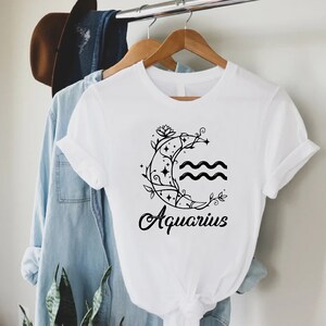 May include: White t-shirt with a black and white graphic of a crescent moon with a vine and stars around it. The text "Aquarius" is below the moon.