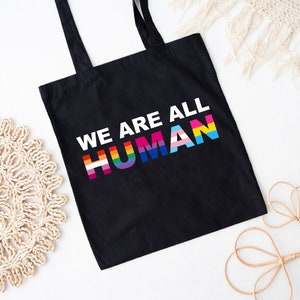 May include: Black tote bag with the text "We Are All Human" in white letters. The word "Human" is in rainbow colors, representing the LGBTQ+ pride flag.