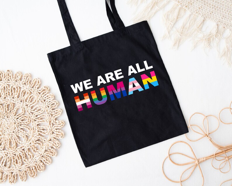 May include: Black tote bag with the text "We Are All Human" in white letters. The word "Human" is in rainbow colors, representing the LGBTQ+ pride flag.