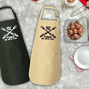 Pu&ograve; includere: Due grembiuli da cucina con un design a tema Star Wars. I grembiuli sono neri e beige con il testo "Che la forchetta sia con te" e una grafica di forchette incrociate.