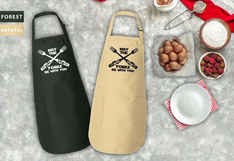 Pu&ograve; includere: Due grembiuli da cucina con un design a tema Star Wars. I grembiuli sono neri e beige con il testo "Che la forchetta sia con te" e una grafica di forchette incrociate.