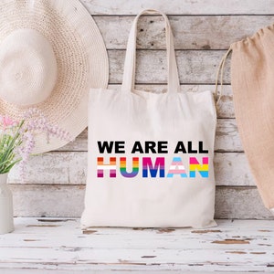 May include: A white canvas tote bag with the text "WE ARE ALL HUMAN" printed in black. The word "HUMAN" is made up of different colored stripes representing the LGBTQ+ pride flag.