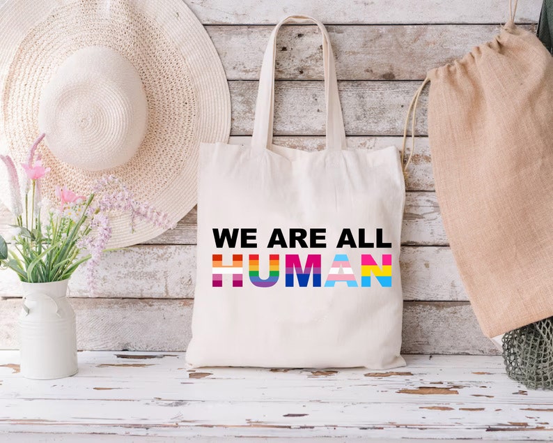 May include: A white canvas tote bag with the text "WE ARE ALL HUMAN" printed in black. The word "HUMAN" is made up of different colored stripes representing the LGBTQ+ pride flag.