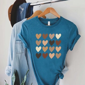 May include: A teal blue t-shirt with a graphic of a grid of 16 hearts in various shades of brown, beige, and cream.