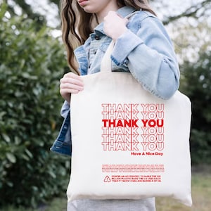May include: White canvas tote bag with red text that says "Thank You" repeated six times and "Have A Nice Day" at the bottom. The bag also has a small recycling symbol and text about the environmental impact of plastic bags.