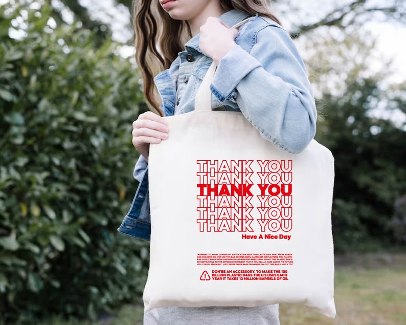 May include: White canvas tote bag with red text that says "Thank You" repeated six times and "Have A Nice Day" at the bottom. The bag also has a small recycling symbol and text about the environmental impact of plastic bags.