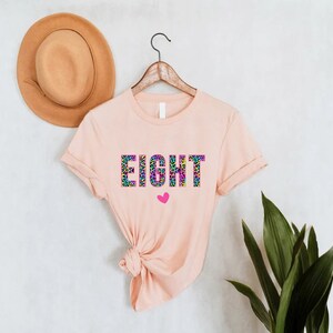May include: A peach-coloured t-shirt with the word "EIGHT" printed in rainbow colours with a leopard print pattern. A small pink heart is below the word.