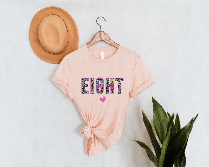 May include: A peach-coloured t-shirt with the word "EIGHT" printed in rainbow colours with a leopard print pattern. A small pink heart is below the word.
